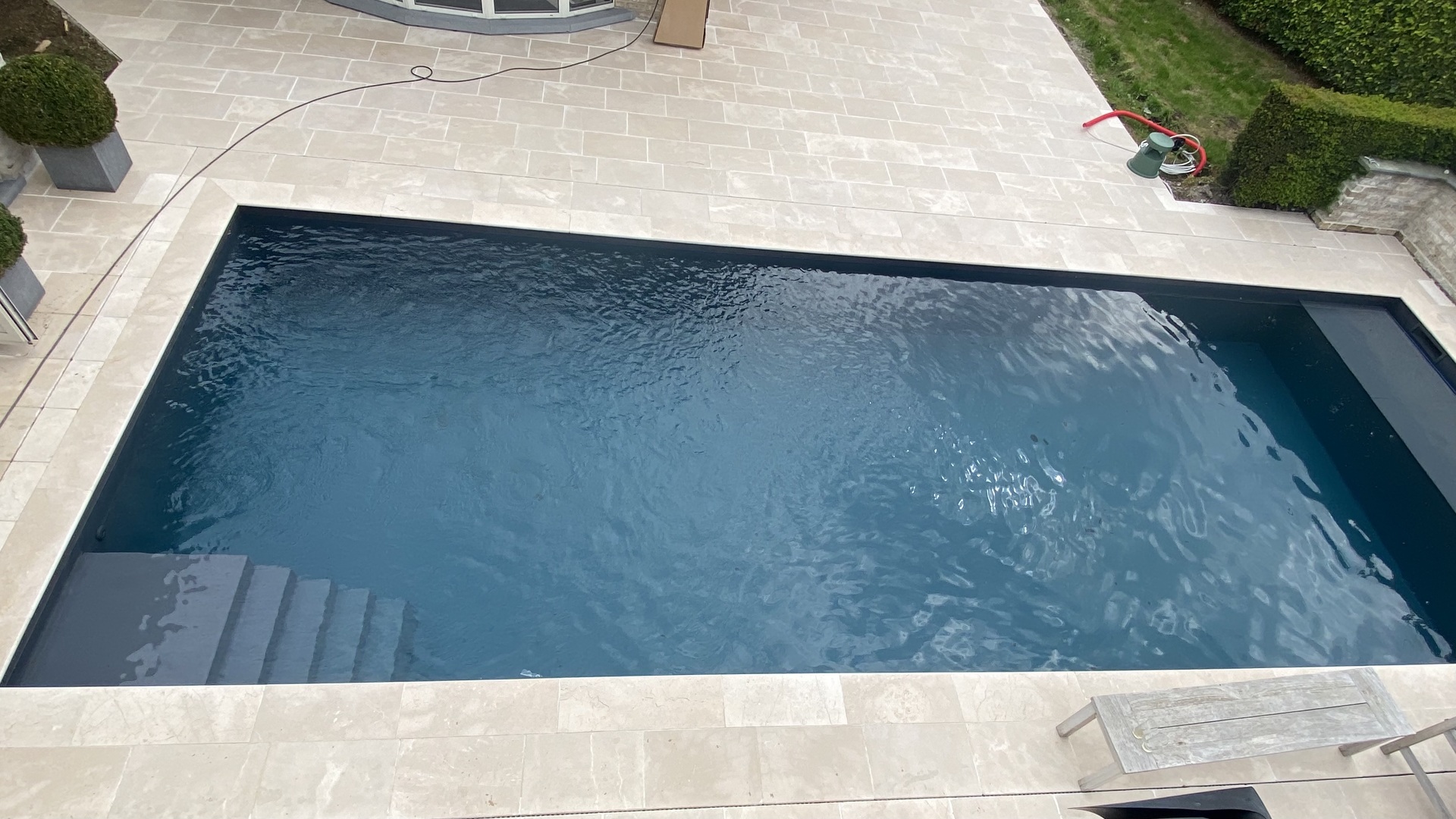 Ingraven zwembad met donkerblauwe liner, strak terras en groene haag op de achtergrond